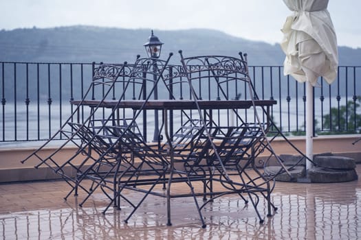closeup detail of black iron metal chairs near wooden table with rain drops on rainy outdoor restaurant terrace