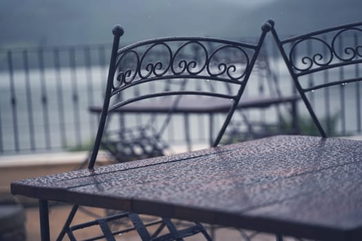 closeup detail of black iron metal chairs near wooden table with rain drops on rainy outdoor restaurant terrace