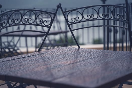 closeup detail of black iron metal chairs near wooden table with rain drops on rainy outdoor restaurant terrace