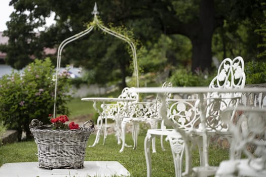 empty green wedding garden with red flowers in wicker basket and white Victorian metal decorative table and chairs
