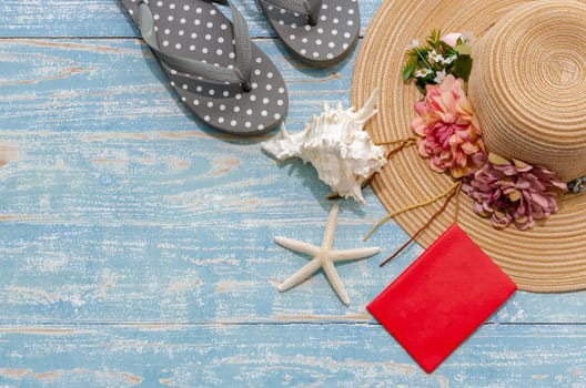 Flat lay summer beach accessories of lady flowered straw hat, flip flops and red passport with seashells and starfish on blue wooden background. Summer beach and holiday trip concept with copy space.