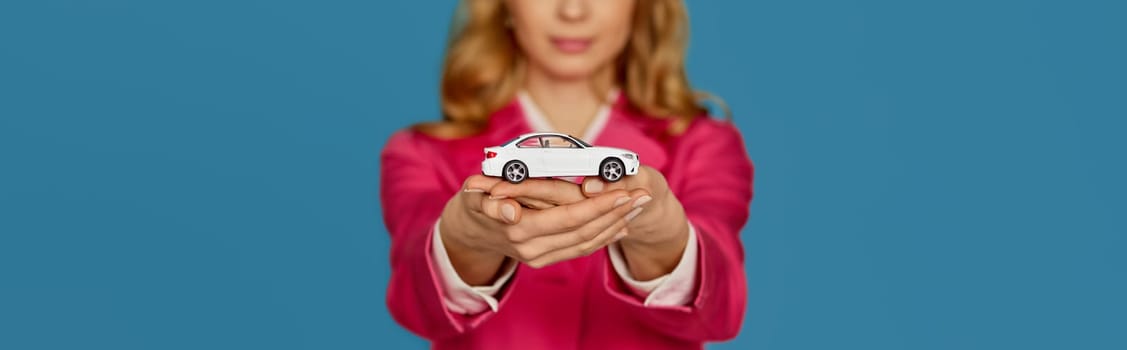 beautiful blonde woman in pink jacket holding little white car on blue background. focus on car. insurance