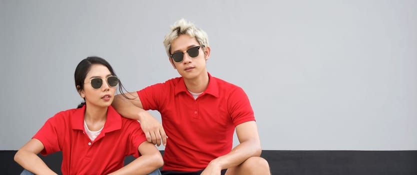 Young asian man in red T-shirt with collar and black sunglasses rest his arm at his girlfriend's shoulder. Portrait on gray background.