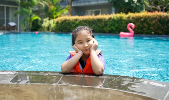 Asian child playing in the pool. Wearing orange life jacket, rests her chin on both hands by the edge of the pool.