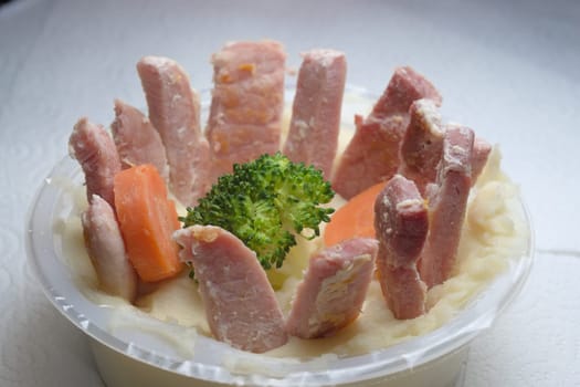 Softly lit funny meal of gammon slices arranged in a circle, on top of a potato mash bed. In the middle is a broccoli floret and two carrot slices