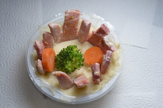Tub of potato mash on top of white kitchen roll. Unusual presentation of meat and vegetables. Homemade twist on a supermarket ready meal