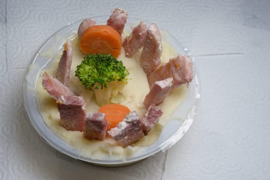 Artistic british cuisine, on a plain white kitchen tissue background. Presented in a round plastic tub with colorful veg and a ring of gammon slices