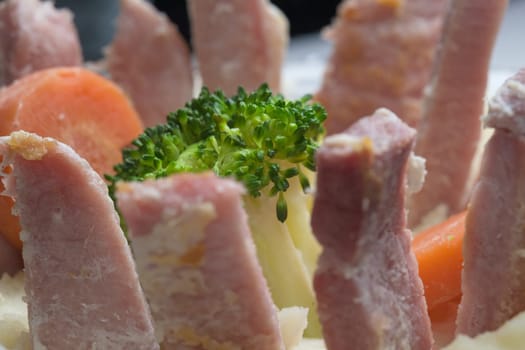 Side view of weirdly presented evening meal. Gammon strips and slices stood upright in white potato mash, focus on broccoli with carrots too.
