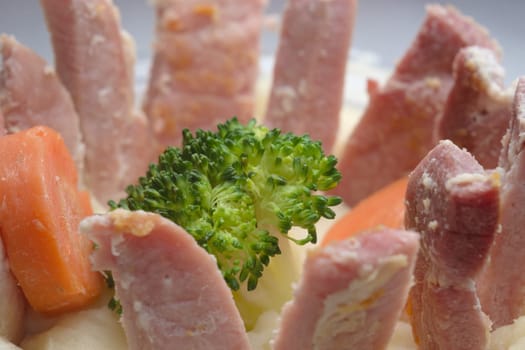Close up image of a weird dinner. A ring of gammon slices embedded in potato mash surround a single broccoli floret and two carrot pieces.