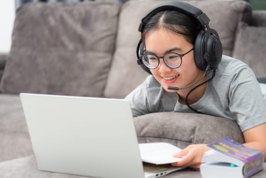 Asian young woman student with headphones lying on the sofa smile look at laptop online learning from school a girl is happy to university internet distance study online work on computer from home