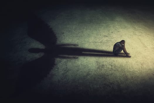 Depressed young man sitting alone isolated in a dark room, casting a shadow of a powerful guardian angel. Inner strength conceptual background. Stressed person introvert with an imaginary friend