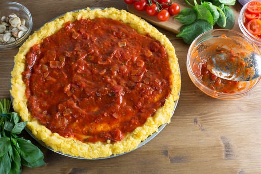 Baking polenta pizza, pizza crust with tomato sauce and empty tomato sauce bowl with spoon on the right side, other ingredients surrounding pizza.