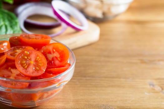 Glass bowl of sliced cherry tomatoes with other ingredients including red onion slices, all to the left side of image with wooden table top to right providing copy space.  Horizontal image.