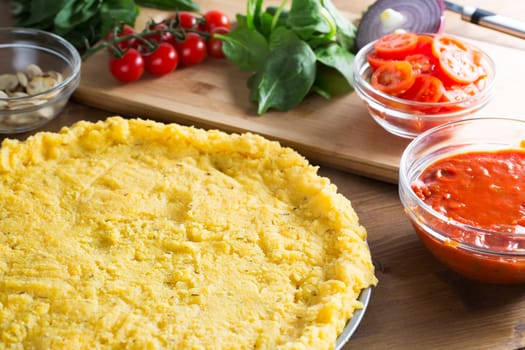 Baking polenta pizza, polenta pizza crust ready for toppings with bowl of tomato sauce to the side and tomatoes, spinach and mushrooms next to pizza crust.
