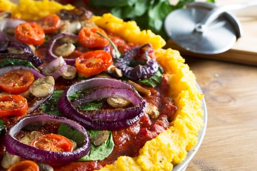 Whole vegan polenta pizza with cherry tomatoes, mushrooms, spinach and red onions.  Close up with pizza slicer out of focus in the background, horizontal orientation.