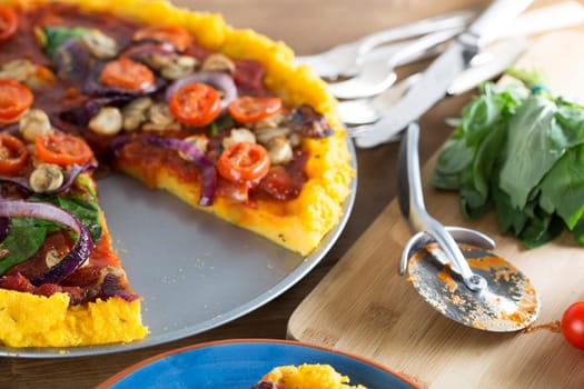 Vegan polenta pizza with cherry tomatoes, mushrooms, spinach and onions with slice removed and pizza slicer to the side.