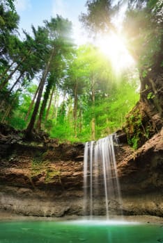 Waterfall on rock layers,above it a mixed forest with blue sky and sun