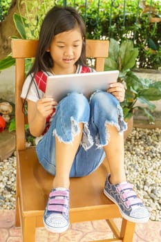 The girl sitting on wooden chair with playing tablet pc.