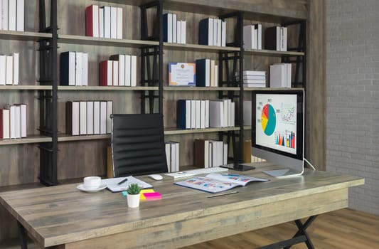 Brown wooden desk with desktop PC and black chair with bookshelves on the wall in modern office room