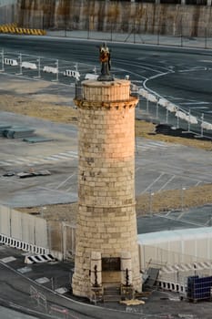 A historic stone watchtower with a statue on top, standing in the port of Civitavecchia. The old maritime structure is surrounded by a modern industrial area. Concept: heritage and travel