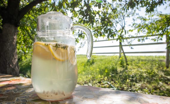 Homemade lemonade made from lemons in a large glass jug on the table in the garden. A jug with lemon and mint stands on the street against the backdrop of greenery on a hot summer day
