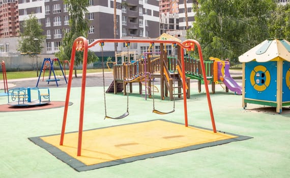 A colorful modern playground in the courtyard in a park without people. Swing, outdoor carousel for children. A playground in a public park surrounded by green trees and new houses. Children exercise