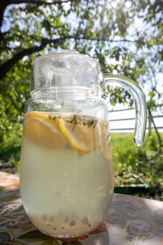 Homemade lemonade made from lemons in a large glass jug on the table in the garden. A jug with lemon and mint stands on the street against the backdrop of greenery on a hot summer day