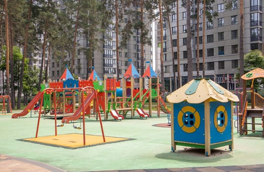 A colorful modern playground in the courtyard in a park without people. Swing, outdoor carousel for children. A playground in a public park surrounded by green trees and new houses. Children exercise