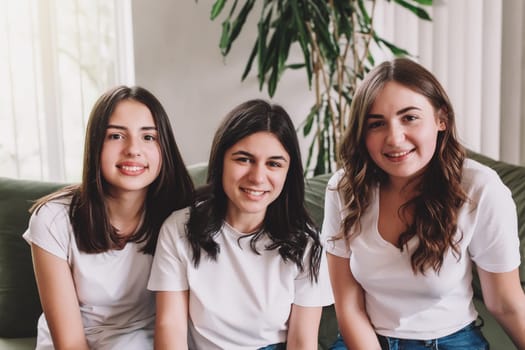 Three pretty smiling caucasian sister girls in white shirt sitting on the green sofa. Friends are posing photo shoot and making a beautiful smile to camera