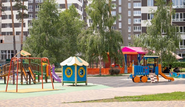 A colorful modern playground in the courtyard in a park without people. Swing, outdoor carousel for children. A playground in a public park surrounded by green trees and new houses. Children exercise