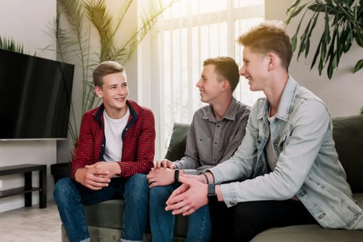 Three male friends are talking at the green sofa in living room. Guys having a talk, sharing and discussing news, cheeling together. Gossip teens