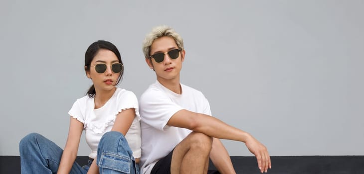 Young asian man in white T-shirt and black sunglasses rest his arm at his knee. His girlfriend sitting next to him. Portrait on gray background.