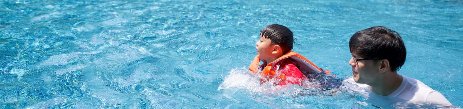 An Asian father teaches his son to swim in the pool. The boy wearing orange life jacket.
