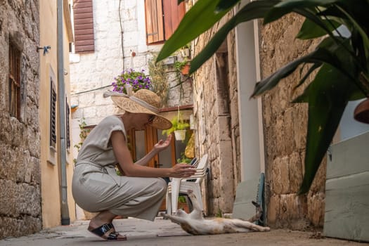Beautiful blonde young female traveler wearing straw sun hat caressing cat while enjoying summer vacation in an old traditional costal town at Adriatic cost, Croatia.