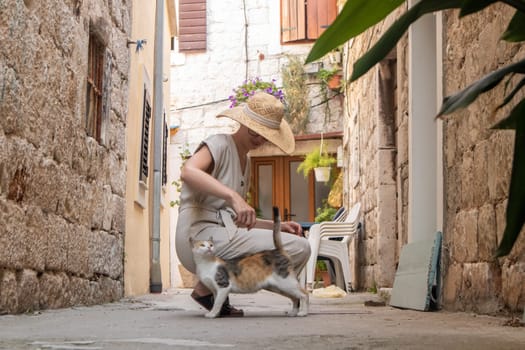 Beautiful blonde young female traveler wearing straw sun hat caressing cat while enjoying summer vacation in an old traditional costal town at Adriatic cost, Croatia.