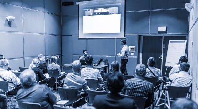 Speaker giving a talk in conference hall at business event. Unrecognizable people in audience at conference hall. Business and Entrepreneurship concept. Blue toned black and white.