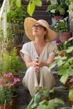 Beautiful female tourist wearing big straw sun hat and sundress sitting and relaxing on old stone house stairs during summer travel on Mediterranean cost on hot summer day
