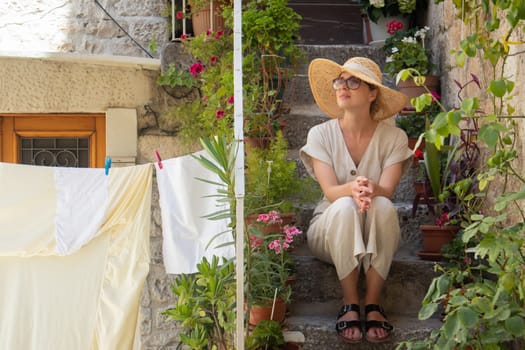Beautiful female tourist wearing big straw sun hat and sundress sitting and relaxing on old stone house stairs during summer travel on Mediterranean cost on hot summer day