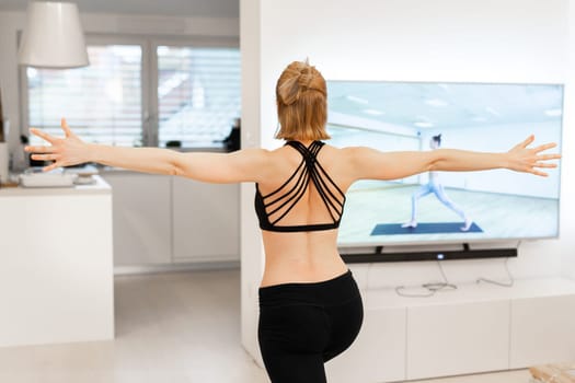 Beautiful blonde woman doing home workout indoors. Woman practice yoga at home. Fit girl using workout tutorials for healthy active lifestyle. Woman using quarantine for home workouts and stretching
