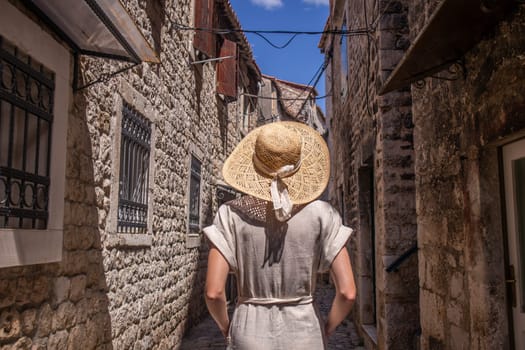 Beautiful blonde young female traveler wearing straw sun hat sightseeing and enjoying summer vacation in an old traditional costal town at Adriatic cost, Croatia. Rear view against the old stone town.