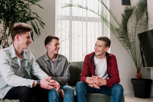 Three male friends are talking at the green sofa in living room. Guys having a talk, sharing and discussing news, cheeling together. Gossip teens