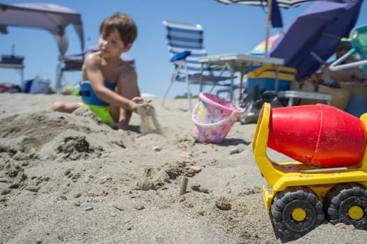 Little boy playing with sand and beach toys. Beach site full of beach furniture