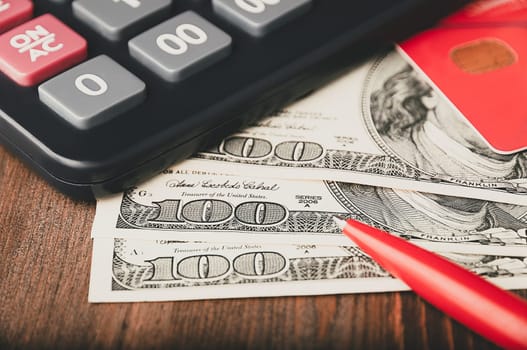 Dollars in one-hundred-dollar bills are spread out on the table next to a calculator, a credit card, and a pen. One-hundred-dollar bills. US dollars. Color style. Layout, template, mockup.