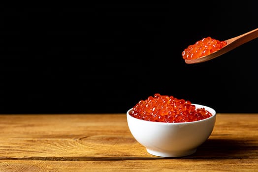 Red caviar in a wooden cup on a black background with a spoon. A large pile of bright caviar. Fresh delicious caviar. A place for advertising, logo, label, layout, mockup. Layout, template.