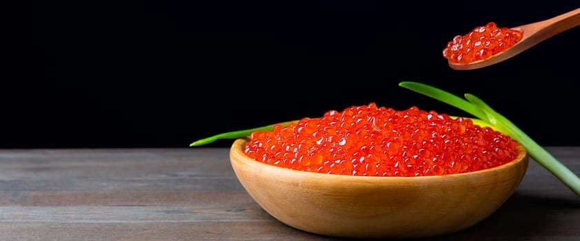 Red caviar in a wooden cup on a black background with a spoon and green onions. A large pile of bright caviar. Fresh delicious caviar. A place for advertising, logo, label, layout, mockup. Layout, template.