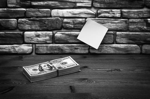 Dollars in one-hundred-dollar bills are stacked on a table near the brick wall. Dollars in a big pile. US dollars. On the brown brick wall there is a place for an inscription, a logo. Black and white style. Layout, template, mockup.
