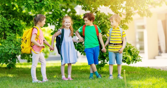 Cute children with rucksacks standing in the park near the school. Concept of elementary studying and friendship.