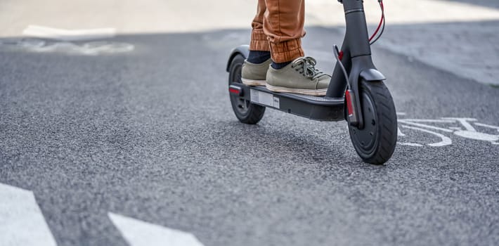 Young man in casual trousers riding an electric scooter detail on his feet and wheel over asphalt road.