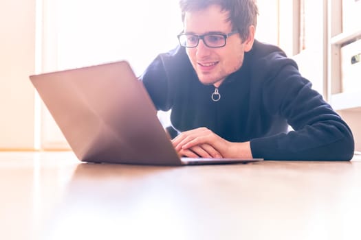 Happy young man with a laptop is working on the wooden floor, light comes in from the window