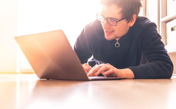 Happy young man with a laptop is working on the wooden floor, light comes in from the window